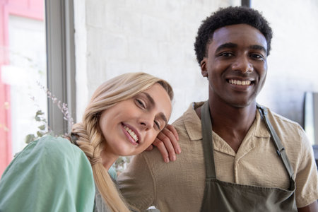 Diverse barista coworkers standing by window with potted plant in cafe wearing aprons and smiling....