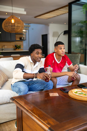 Diverse male friends sitting on sofa in modern living room, holding soda bottles and eating pizza....