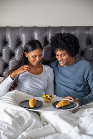Diverse couple leaning against headboard on bed sharing breakfast tray with croissants and fruit....