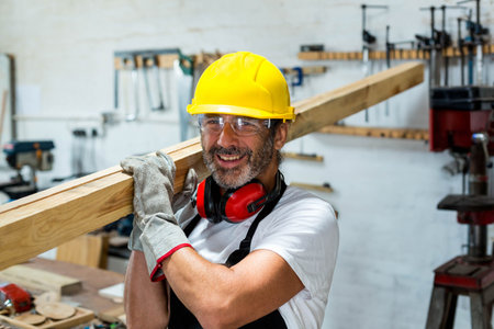 Mature male tradesman holding long wooden beam on shoulder in woodshop, wearing yellow hard hat....