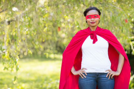 African American woman posing hands-on-hips in sunny park under trees in red cape mask, copy space....