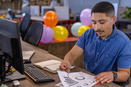Man in thirties sitting at open office desk with balloons reviewing charts with pen, copy space....