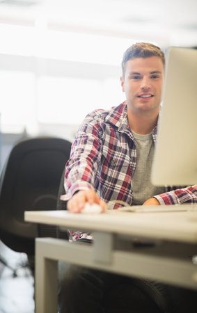 Twenty-something man wearing red plaid shirt sitting at desk in bright lab using mouse and monitor....