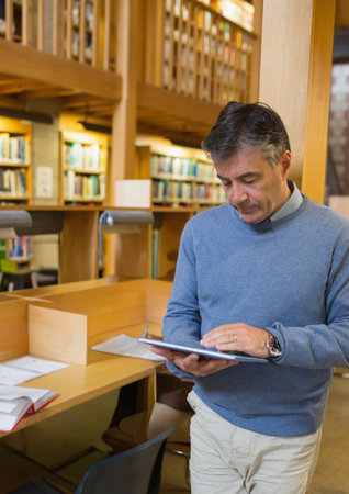 Man wearing sweater standing by wooden carrel using tablet with open notebook, copy space. Library,...