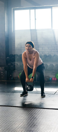 African American woman performing kettlebell hinge in workout clothes in warehouse gym. Industrial,...