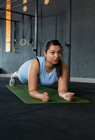 Female in light blue sportswear holding forearm plank on green mat at gym with rings. Fitness,...