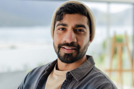 Indian man posing by window with waterfront, wearing beanie T-shirt and overshirt blurred. Portrait,...