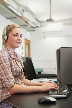 Adult female sitting at computer lab wearing plaid shirt and headset, typing at keyboard and mouse....