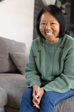 Senior Asian woman sitting on grey sofa at home with cushions, pillow, plant, copy space. Senior,...