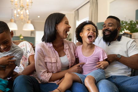 Diverse family laughing together on sofa in living room wearing headphones under chandelier. Family,...