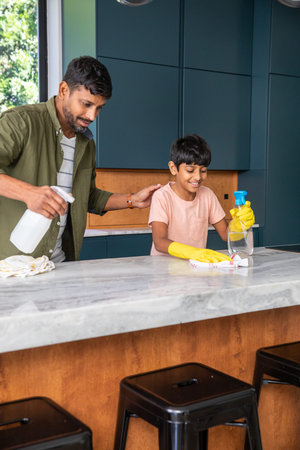 Indian father and son cleaning marble countertop in modern kitchen using spray bottles and cloth....