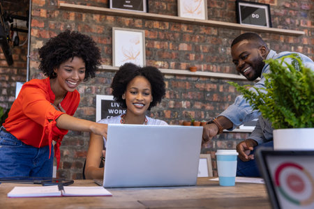 African American coworkers collaborating around laptop at desk in modern workspace with coffee cup....