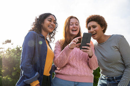 Diverse female friends standing together in sunny park laughing while looking at smartphone. Casual,...