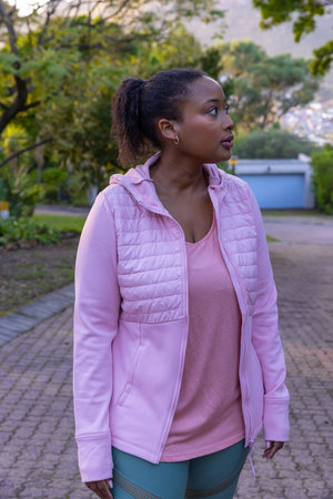 Mid adult African American woman standing on brick path wearing pink activewear and hoop earrings....