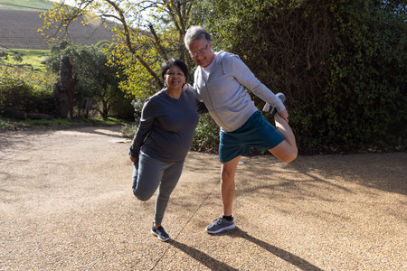 Diverse senior couple in athletic wear performing quadriceps stretch on sunlit gravel driveway....