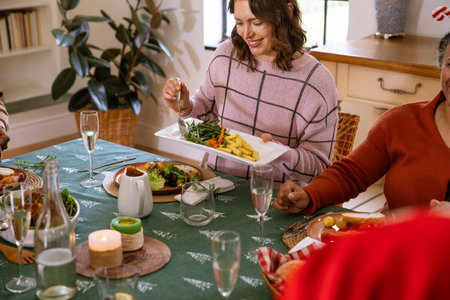 Diverse female family passing vegetable platter at home dining table with candlelight. Festive,...
