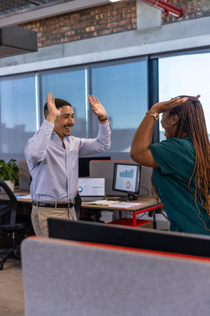Diverse coworkers in business attire high-fiving at open-plan office with computer monitors....