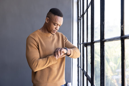 Mid adult African American man checking smartwatch beside metal-framed window in office, copy space....