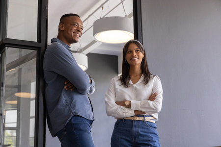 Diverse coworkers leaning against glass partition in office corridor under pendant lamps....