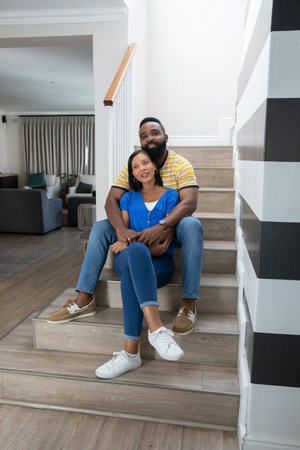 African American couple sitting on wooden staircase embracing each other in home foyer with sofa....