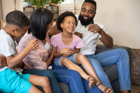 Diverse family sitting on gray sofa in living room, father tickling daughter and mother watching....