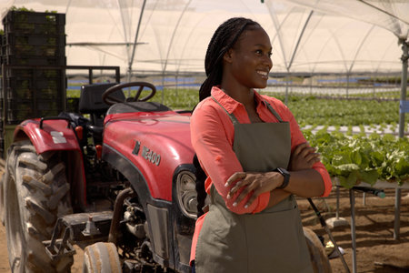African American woman wearing green apron posing in greenhouse alongside red tractor and crates....