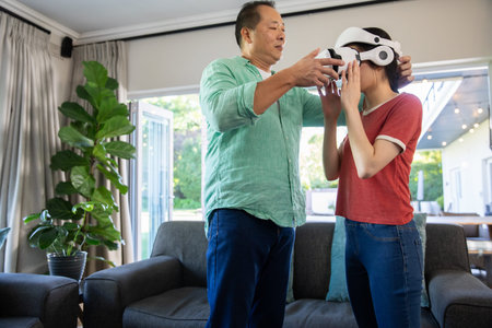 Asian father and daughter helping put on white virtual reality headset in living room. Family,...