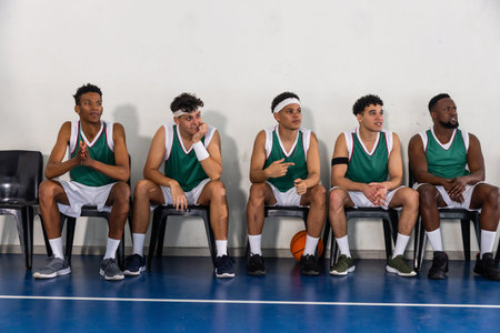 Diverse male basketball teammates resting on chairs at gym wearing green uniforms with basketball....