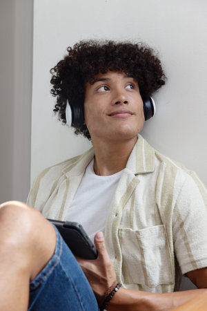 African American male sitting on floor at home wearing headphones and holding smartphone. Casual,...