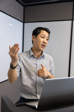 Asian man presenting behind podium in meeting room with laptop computer and frosted glass panels....
