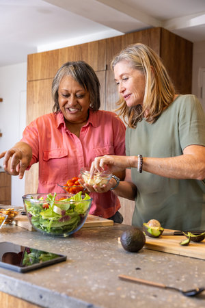 Diverse senior friends preparing salad on kitchen island with glass salad bowl and tablet. Culinary,...