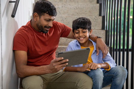 Indian father and son sitting on staircase landing at home viewing tablet computer together. Family, bonding, technology, candid, lifestyle, modern, educationの写真素材