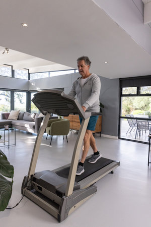 Senior man walking on treadmill at home wearing sportswear with sliding glass doors and armchair....