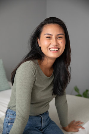 Asian woman sitting on sofa in bedroom wearing olive green top and jeans with cushion, plant....