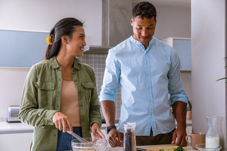 Diverse couple stirring batter in glass bowl, chopping herbs on cutting board at kitchen counter....