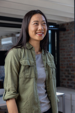 Asian woman standing in office under exposed beams by desk and air conditioner near brick wall....