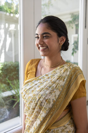 Indian woman wearing yellow sari beside large window at home smiling, gazing through green foliage. Botanical, serenity, elegance, natural light, interior design, cultural attire, wellnessの写真素材