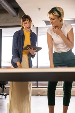 Female coworkers collaborating at office desk while examining tablet and sticky notes. Collaboration, teamwork, innovation, modern, professional, workspace, productivityの写真素材