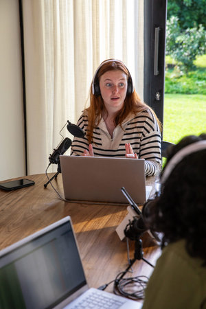 Colleagues recording podcast at wooden table next to garden door with laptop and microphone. Studio, communication, creativity, broadcast, electronics, collaboration, outdoorsの写真素材