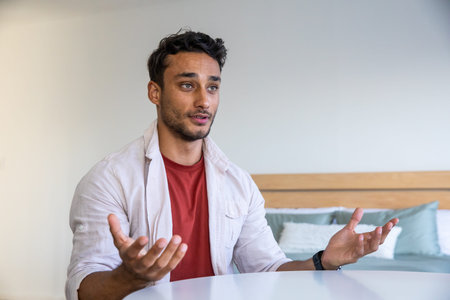 Man in twenties sitting at guest room white table speaking to camera with pillows on headboard. Casual, conversation, modern, comfort, communication, relaxed, lifestyleの写真素材