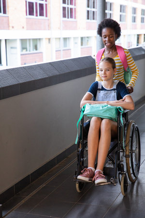 Diverse teenage female classmates pushing wheelchair student along campus walkway beside brick wall. Inclusive, friendship, accessibility, school, urban, youthful, teamworkの写真素材