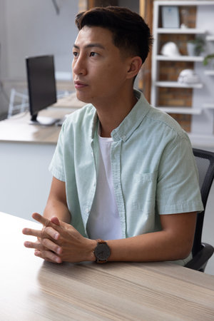 Asian man wearing mint-green shirt sitting at desk in office looking toward shelving. Professional, workspace, minimalism, interior, contemporary, productivity, corporateの写真素材