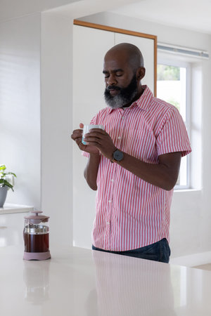 Senior African American man holding ceramic mug smelling coffee near French press on kitchen island. Maturity, relaxation, wellness, cozy, contemporary, lifestyle, tranquilityの写真素材