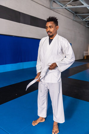 African American man standing in white gi adjusting white belt on interlocking mats inside dojo. Martial arts, training, fitness, discipline, dojo, athleticism, focusの写真素材
