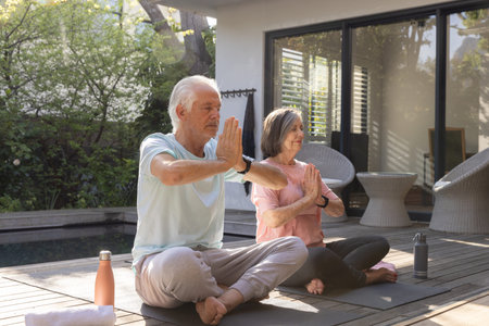 Senior couple practicing yoga on yoga mats on wooden deck beside pool with reusable water bottles. Wellness, tranquility, outdoor, relaxation, harmony, nature, mindfulnessの写真素材