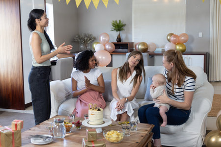 Diverse female friends gathering around coffee table at home celebrating first birthday with cake. Celebration, friendship, family, joy, warmth, domestic, colorfulの写真素材