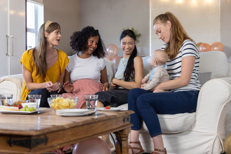 Diverse female friends sitting on upholstered couch in living room with balloons holding infant. Celebration, friendship, family, cozy, elegant, intimate, joyの写真素材