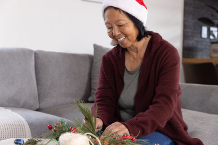 Senior Asian woman with Santa hat arranging pine branches, berries into centerpiece on coffee table. Festive, seasonal, cozy, handcrafted, rustic, joyful, traditionalの写真素材