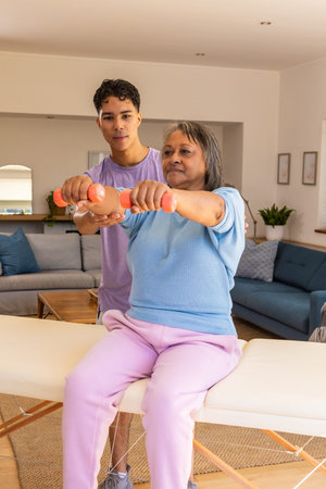 Senior woman client and trainer lifting orange dumbbells on bench in living room guiding posture. Fitness, wellness, training, rehabilitation, active aging, interior, healthの写真素材