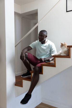 African American man sitting on wooden stairs at home petting cat wearing wristwatch. Minimalist, cozy, relaxed, lifestyle, contemporary, warm, interiorの写真素材